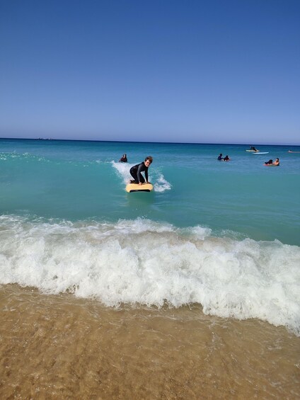 A person surfing on a yellow board in the ocean with clear blue water and a sandy beach in the foreground. 2