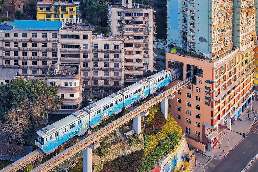 A blue train on an elevated track passing through a cityscape with multi-story buildings and greenery. 1