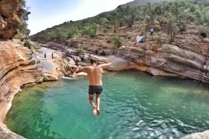 A man jumping into a natural pool surrounded by rocky terrain and sparse vegetation. 1