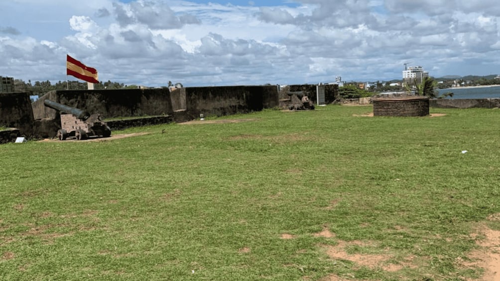 A grassy field with a flag, a cannon, and a building in the background. 2