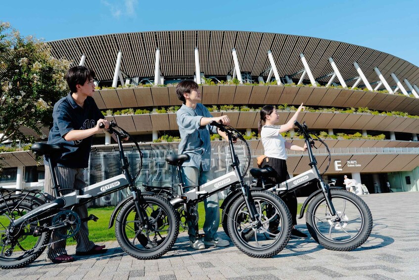 Three people with electric bicycles in front of a modern building with a curved roof and vertical slats. 1
