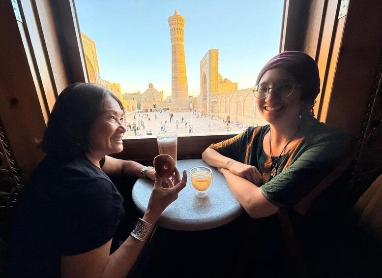 Two people sitting at a table with drinks, overlooking a historic square with a prominent tower. 1