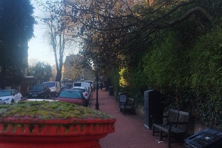 A narrow street lined with parked cars, a red bench, and a large planter. 2