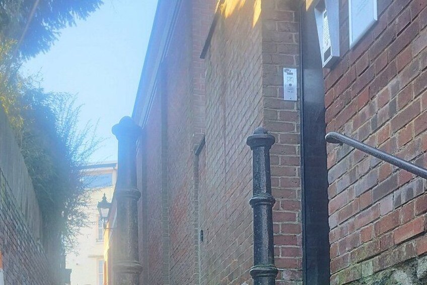 A narrow alleyway with brick buildings, a metal railing, and a street lamp. 4