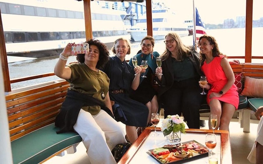 A group of people on a boat, sitting on benches and holding drinks, with a table of food and drinks in front of them. 5