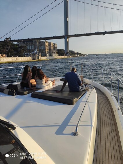 A boat with people relaxing on the deck, a bridge in the background, and a cityscape across the water. 1