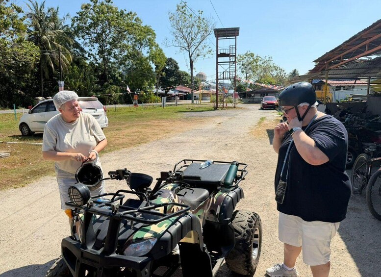 Two people standing next to an all-terrain vehicle on a dirt road with cars and buildings in the background. 3