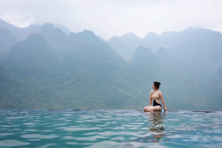 A person sitting on the edge of a clear blue pool with mountains in the background. 4