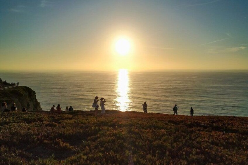 A sunset over the ocean with people on a cliff and a grassy area in the foreground. 2