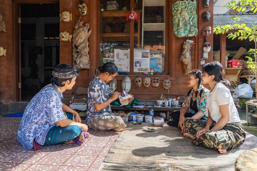 A group of people sitting on the ground in front of a shop displaying various masks. 1