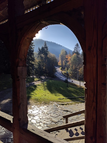 A wooden gazebo with a stone-tiled floor, overlooking a forested hillside with a road and a mountain in the background. 3