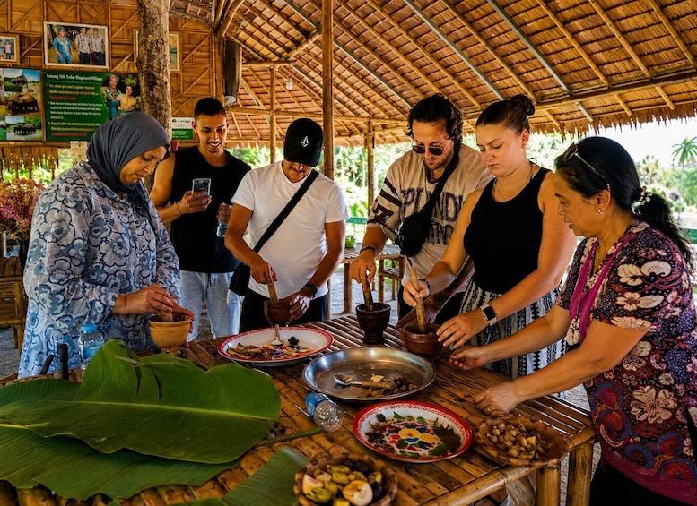 A group of people preparing food outdoors at a rustic wooden table with a thatched roof, surrounded by lush greenery. 5