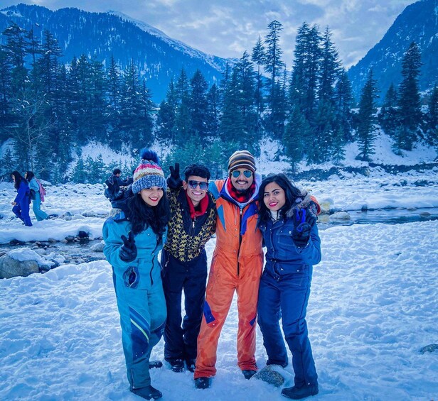 A group of people in winter clothing posing in a snowy mountainous area with a river and trees in the background. 2