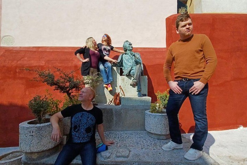 A man in a brown sweater and jeans standing on a stone platform with a statue and people in the background. 3