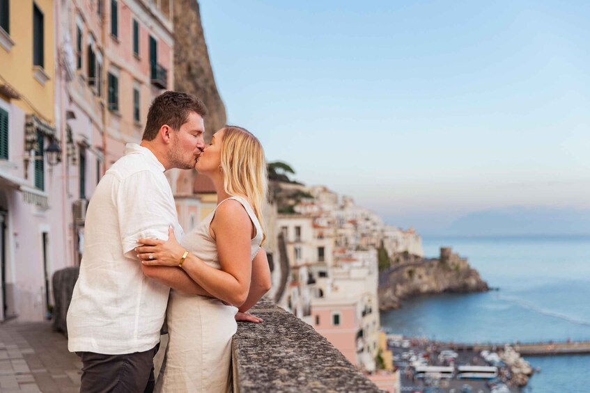 A couple embracing on a stone ledge overlooking a coastal town with colorful buildings and a cliff. 1