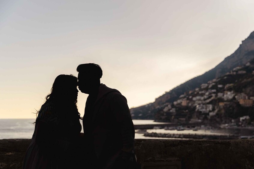 A couple embracing against a coastal backdrop with a mountain and buildings. 2