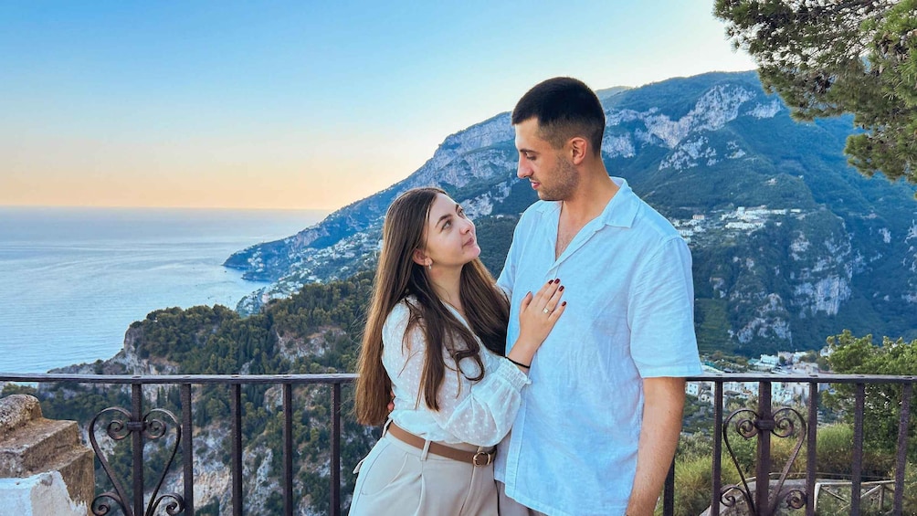 A couple standing on a balcony overlooking a coastal landscape with mountains and the sea. 4