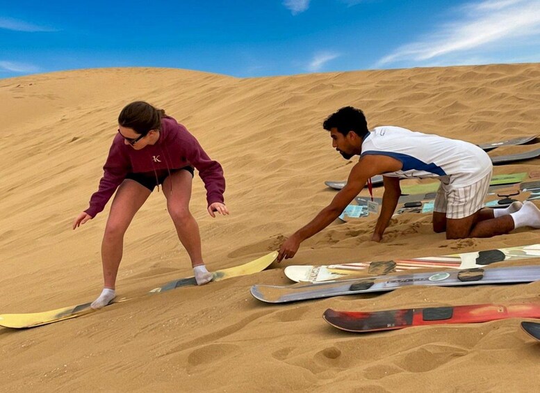 A person in a purple jacket and another in a white shirt on a sandy slope with skis and snowboards. 1