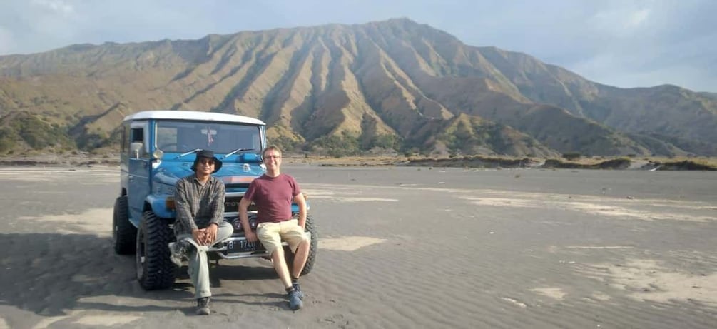 A blue vehicle with two people sitting in front, a desert landscape with mountains in the background. 4
