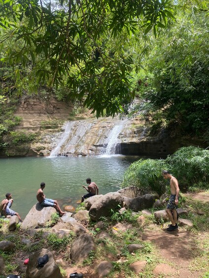 A waterfall, people sitting on rocks, and a lush green environment. 1