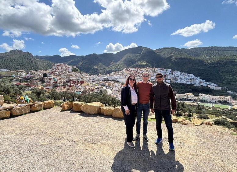 Three people standing on a rocky outcrop with a coastal town and mountains in the background. 5