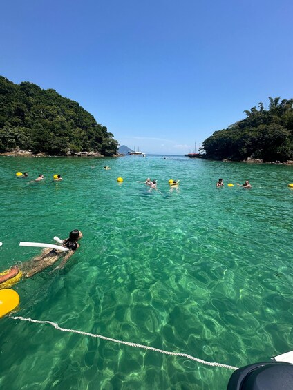 People swimming in clear turquoise water with yellow buoys, surrounded by lush green hills. 3