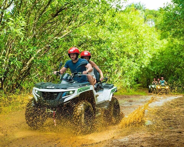 Two people on an ATV in a muddy path surrounded by dense greenery. 3