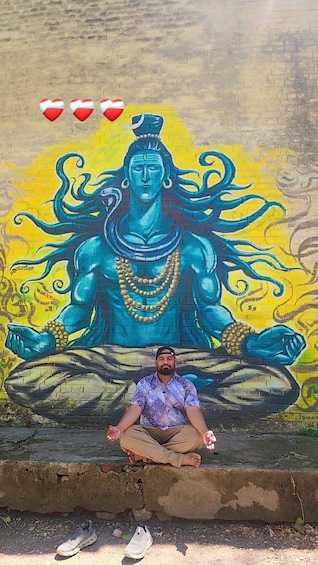 A man sitting in a meditative pose in front of a mural of a deity. 2