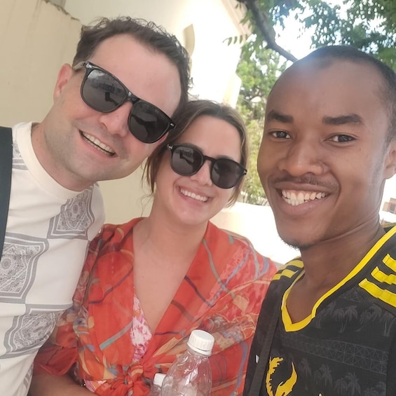 Three people posing together outdoors, with one person wearing a patterned shirt, another in a floral dress, and the third in a sports jersey. 2