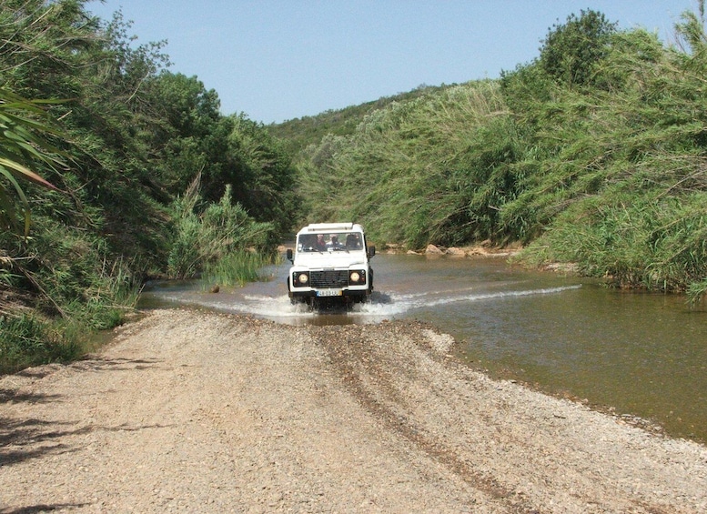 A white off-road vehicle driving through a shallow river on a gravel path surrounded by dense greenery. 3