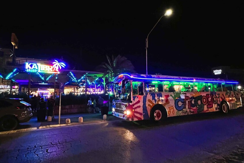 A brightly lit bus with colorful graffiti parked in front of a building with neon signs. 5