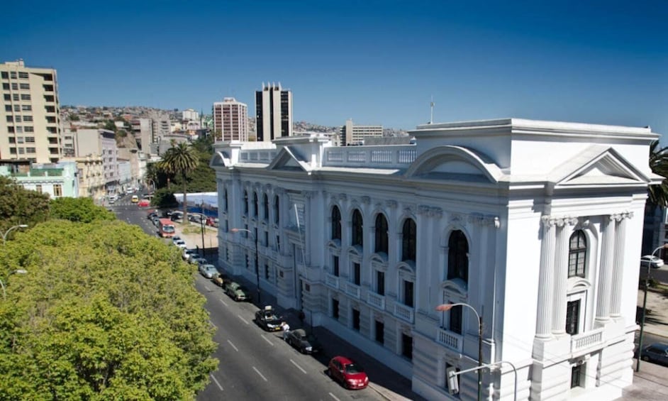 A white classical building with columns and arched windows, surrounded by a cityscape and a clear blue sky. 3