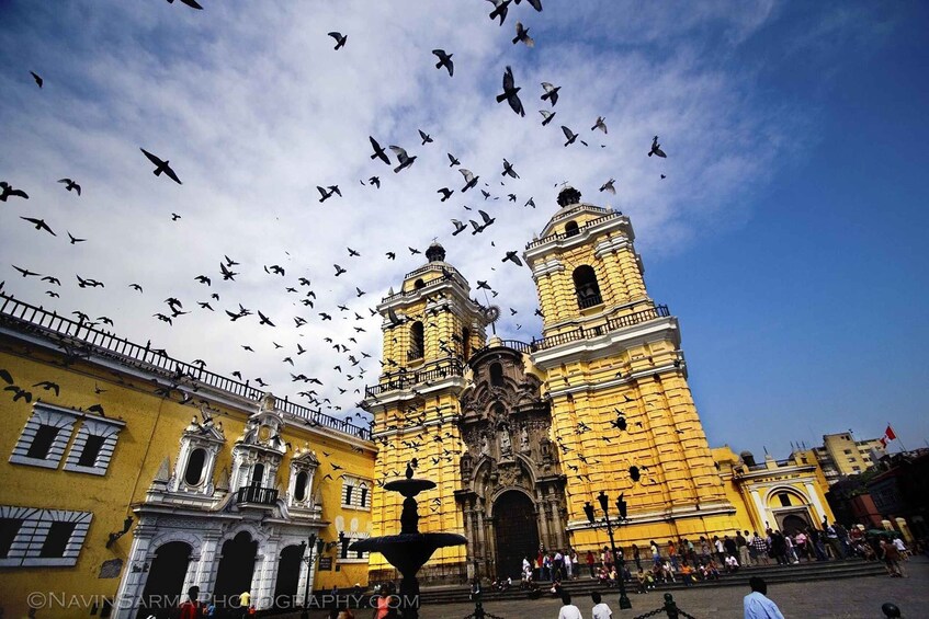 A historic yellow building with two towers and a fountain in front. 1