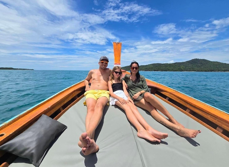 Three people on a boat with a wooden hull and a grey cushion, on a calm sea with a clear sky and a distant green island. 1