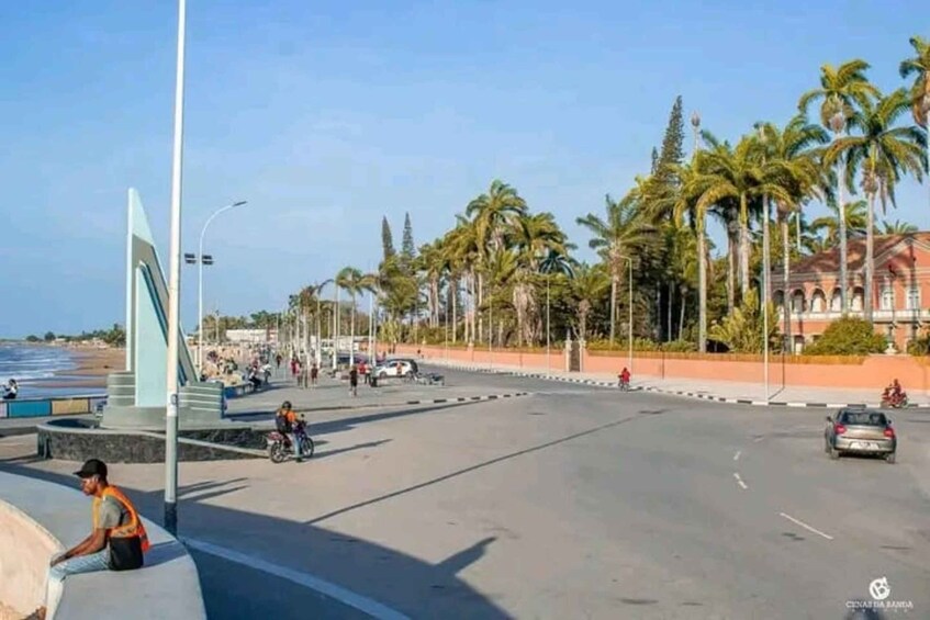 A coastal road with palm trees, a monument, and a beach in the background. 1