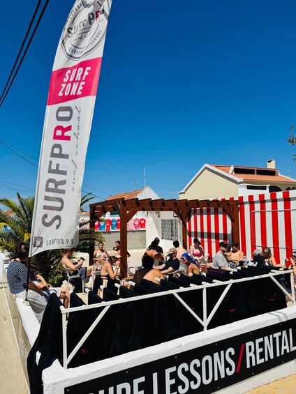 A SURF PRO surf school with a SURFZONE flag, people relaxing, and a rental sign. 3