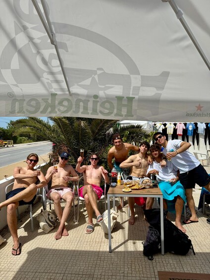 A group of people sitting under a tent with a table in the center, holding drinks and enjoying a sunny day outdoors. 4