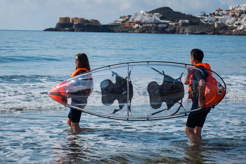 Two people carrying a transparent kayak in shallow water with a coastal town in the background. 4