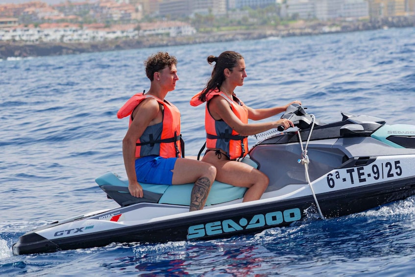 Two people on a Sea-Doo jet ski, wearing life jackets, with a cityscape in the background. 2
