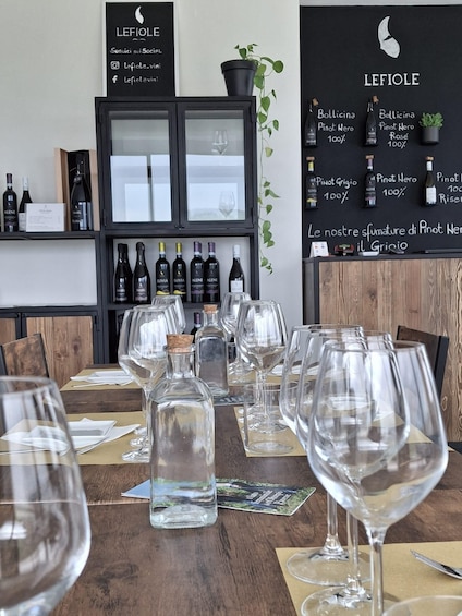 A wine tasting room with empty wine glasses on a wooden table, a selection of wine bottles on shelves, and a chalkboard menu listing wine types. 2