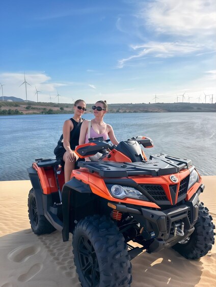 Two people on an orange ATV on a sandy beach with wind turbines in the background. 1