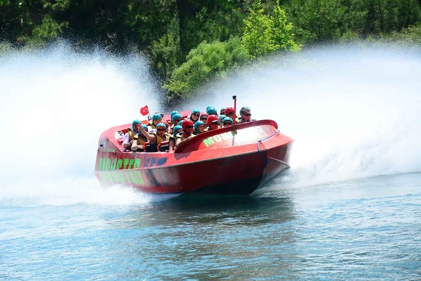A red boat with people wearing helmets and life jackets, moving through water and creating a large spray. 1