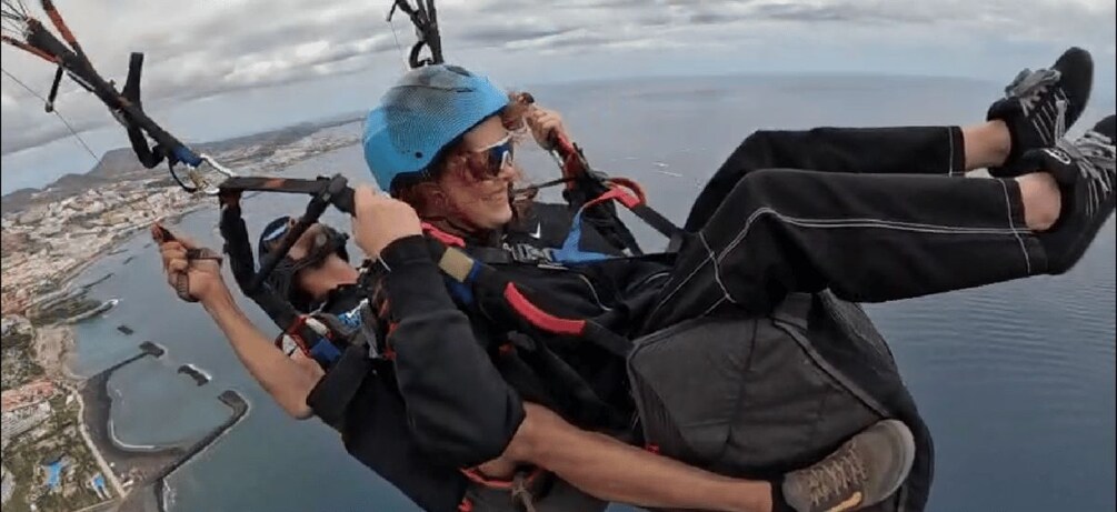 A person skydiving with a parachute, another person holding the harness, and a coastal landscape with water and buildings below. 2
