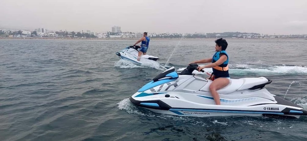 Two people jet skiing on a body of water with a cityscape in the background. 1