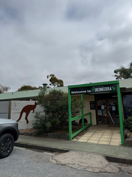A building with a kangaroo silhouette on the wall, a sign reading 'Welcome to URIMBIRRA,' and a parked car. 2