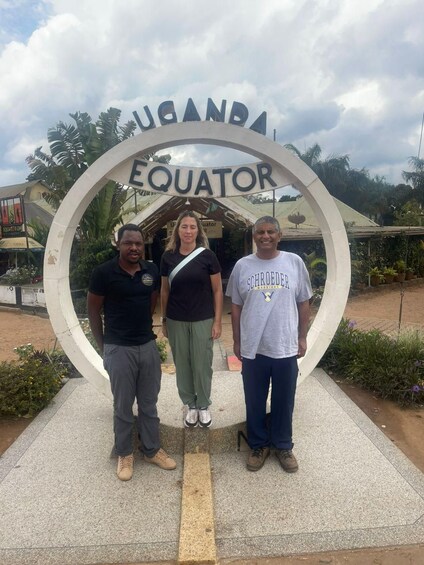 Three people standing in front of a sign that reads 'UGANDA EQUATOR'. 3
