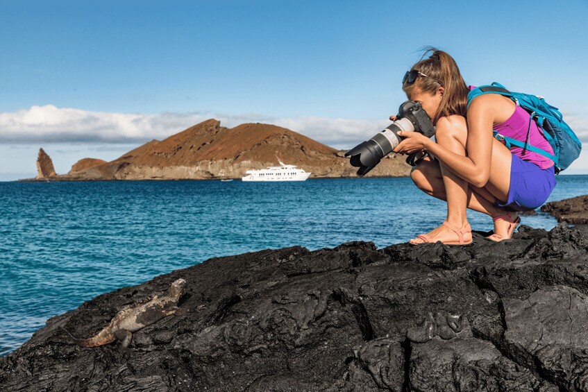 A person crouching on a rocky shore with a camera, a boat in the background, and a mountainous landscape. 2