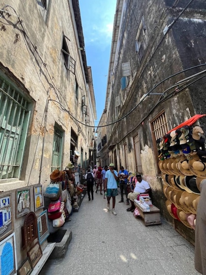 A narrow street with buildings on either side, some with barred windows, and a vendor selling hats. 5