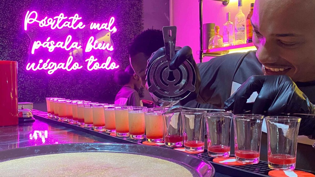 A bar setting with a neon sign, glasses of drinks, and a bartender preparing a drink. 5