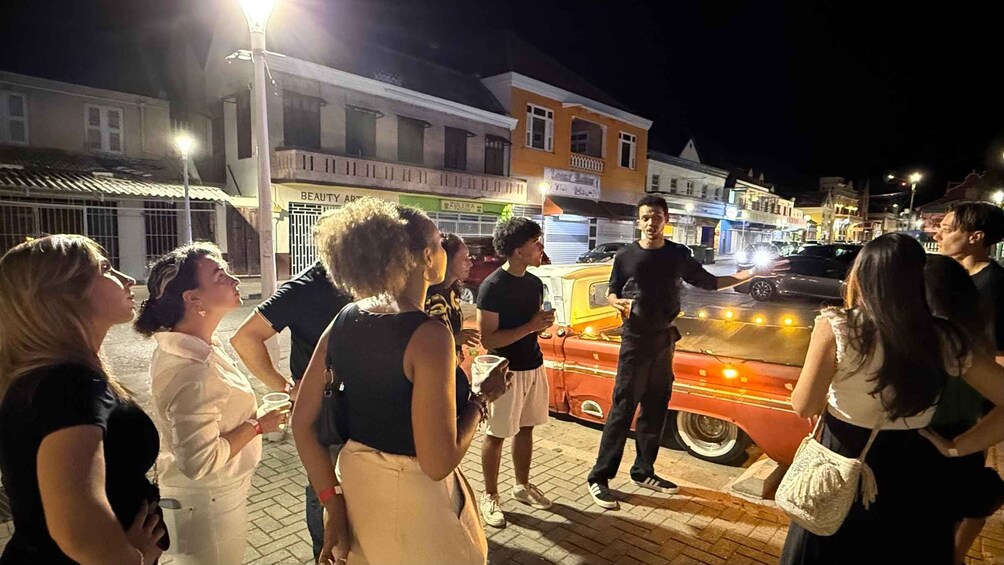 A group of people gathered around a vintage car on a city street at night. 2